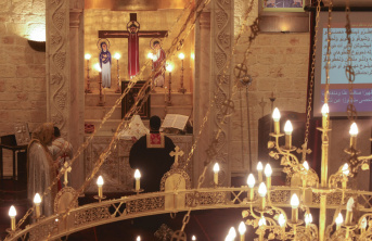Interior of a church with lit candles in the foreground and a priest praying to the cross in the background